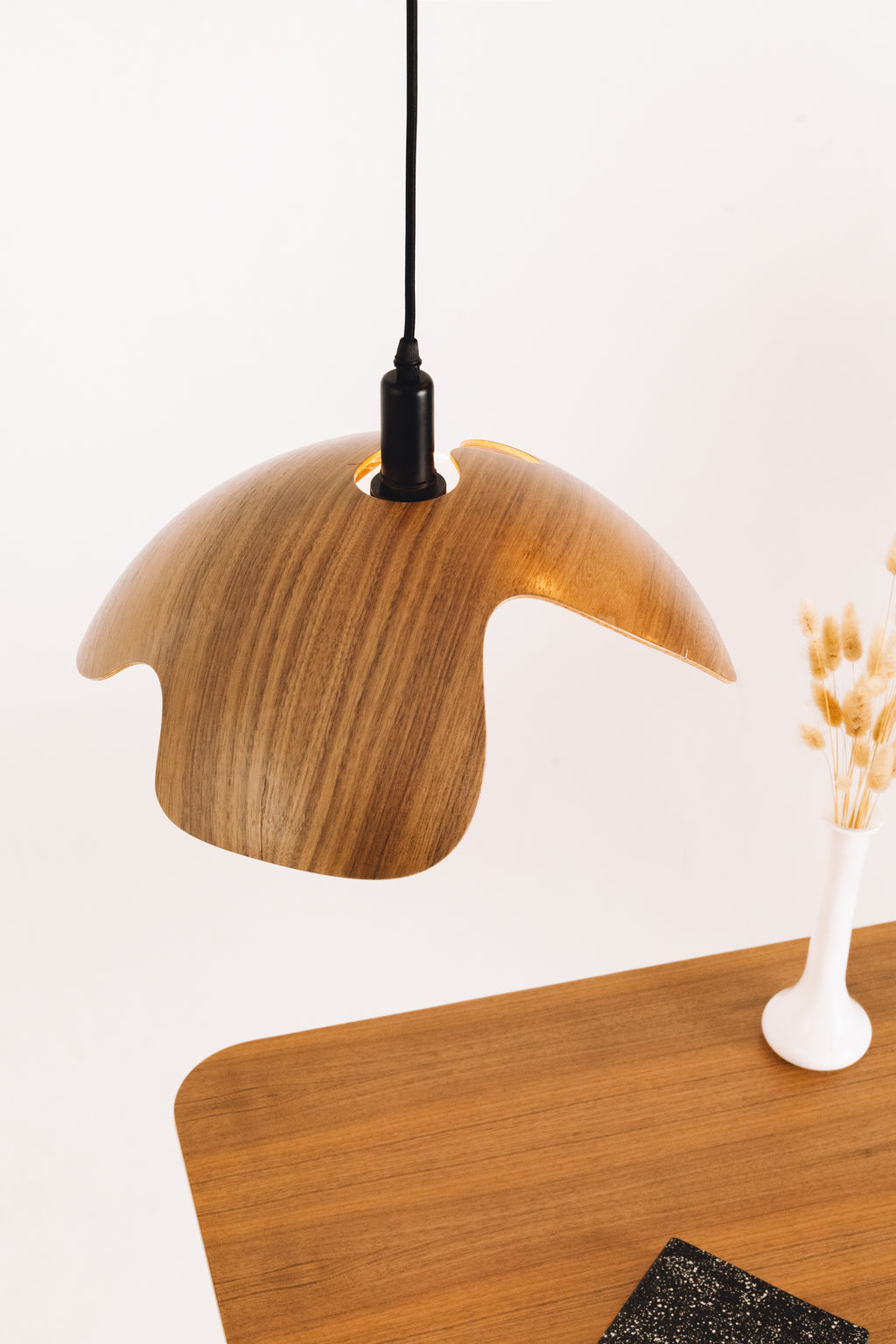 Wooden pendant lamp hanging above a wooden table with a vase of dried flowers on a light background